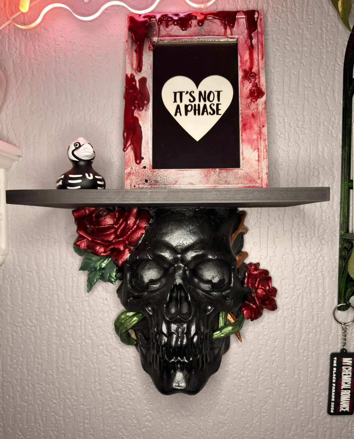 Thorn & Bone gothic skull wall shelf in glossy black with crimson roses, green vines and copper thorns, holding a framed print